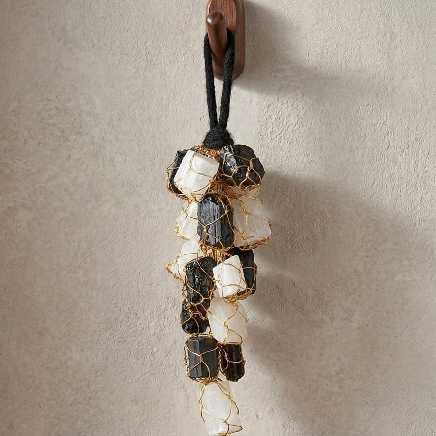 Handcrafted Black Tourmaline and Selenite crystal hanging featuring natural rough stones wrapped in gold wire, suspended from a black cord on a wooden hook against a textured beige wall. Ideal for home décor, energy cleansing, and protection.